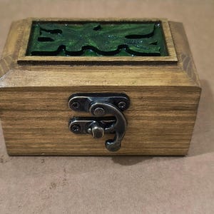 Puede incluir: Una caja rectangular de madera con un acabado marrón natural y una incrustación texturizada de color verde oscuro en la tapa. La parte frontal presenta un mecanismo de cierre de metal plateado fijado con tornillos pequeños.