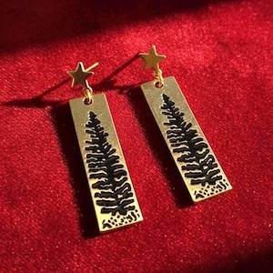 May include: Gold-tone earrings with a rectangular pendant featuring a black tree design. Each earring has a small gold star at the top. The earrings are displayed on a red velvet surface.