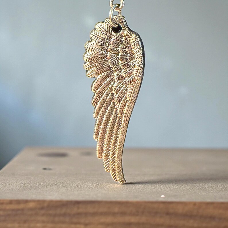 May include: A gold-colored wing pendant with intricate feather detailing. The pendant is suspended from a small loop at the top. The wing is displayed against a blurred background and rests on a wooden surface.
