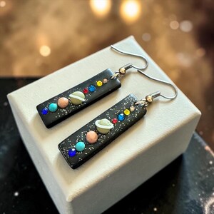 May include: A pair of dangle earrings featuring a black rectangular design with a galaxy theme. Each earring has a series of colorful, spherical beads and a planet-shaped bead. The earrings have silver-colored fish hook ear wires.