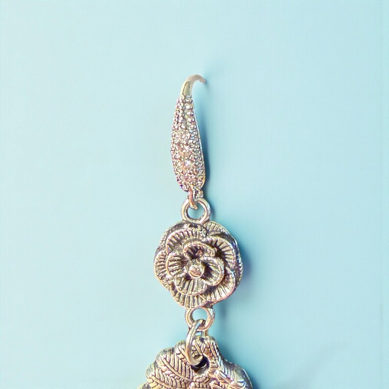 May include: A silver-toned earring with a floral design and a leaf-shaped charm. The earring features a decorative hook and a textured, sparkling upper section. The background is a solid light blue.