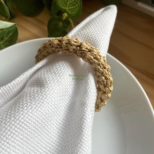 May include: A natural woven napkin ring made of straw, in a light brown color. The ring is placed on a white napkin and a white plate.