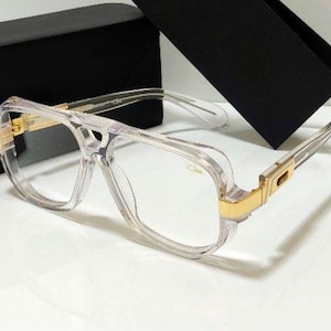 Puede incluir: Gafas transparentes con detalles dorados en la montura y las patillas. Las gafas tienen forma cuadrada y están sobre una superficie blanca con un estuche negro en el fondo.