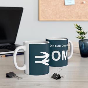 Può includere: Due tazze in ceramica bianca con un design blu scuro. La tazza a sinistra ha una freccia bianca e un simbolo bianco. La tazza a destra ha il testo "Old Oak Common" e "OM" in bianco.