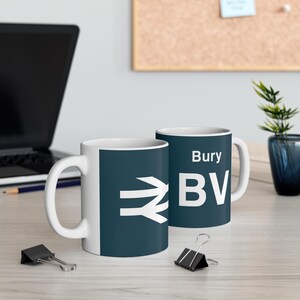 May include: Two white ceramic mugs with a blue and white design. One mug has the text "Bury BV" and the other mug has a stylised train track symbol.