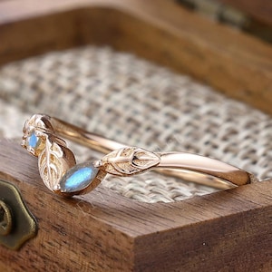 Puede incluir: Un anillo de oro rosa con diseño de hojas y dos piedras preciosas azules. El anillo está en una caja de madera con forro de tela clara. El anillo tiene forma curva, posiblemente una alianza o un anillo de moda.