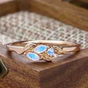 May include: A rose gold ring featuring a leaf motif and three blue gemstones. The ring is displayed in a wooden jewellery box. The gemstones are oval and have a blue tint. The ring's band has a delicate, nature-inspired design.
