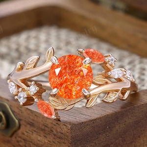 May include: A gold ring featuring an oval, orange gemstone as the centerpiece, accented by smaller orange stones and clear crystals. The ring's band is designed with leaf and vine motifs, set against a wooden box.