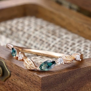 Puede incluir: Un anillo de oro rosa con diseño de hojas, con gemas de color verde azulado y pequeños diamantes blancos. El anillo se exhibe dentro de una caja de madera con un forro de arpillera natural, mostrando los delicados detalles de la joya.