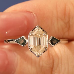May include: A silver ring with a central, elongated, hexagonal-cut diamond. Two smaller, dark green, diamond-shaped stones are on either side of the main stone. The ring is set against a blue background.