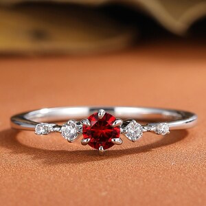 May include: A silver-toned ring featuring a central round-cut red gemstone flanked by two smaller clear stones on each side, set on a thin polished metal band with a delicate prong setting that secures each of the five stones in place.