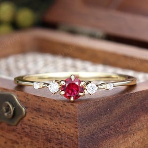 May include: A gold ring featuring a central round-cut red gemstone flanked by two smaller clear stones on each side. The band is thin and polished, showcasing a delicate design that holds the stones in a prong setting for a refined appearance.