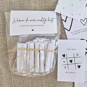 Puede incluir: Una bolsa transparente atada con hilo dorado contiene 30 notas de papel blanco enrolladas. Una tarjeta blanca con el texto "Warum du mir wichtig bist" y otras tarjetas con gráficos y texto en negro están dispersas.