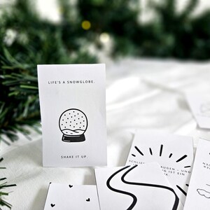 May include: A collection of white cards with black illustrations and text. One card features a snow globe graphic with the text "LIFE'S A SNOWGLOBE. SHAKE IT UP." Other cards have minimalist designs, including a road, sun rays, and clouds.