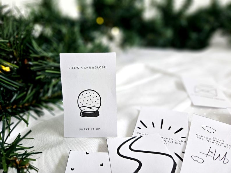May include: A collection of white cards with black illustrations and text. One card features a snow globe graphic with the text "LIFE'S A SNOWGLOBE. SHAKE IT UP." Other cards have minimalist designs, including a road, sun rays, and clouds.