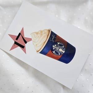 May include: Watercolor illustration of a Starbucks coffee cup with whipped cream, a pink star, and the word "Santa" written on the cup. The cup is brown and blue with the Starbucks logo.