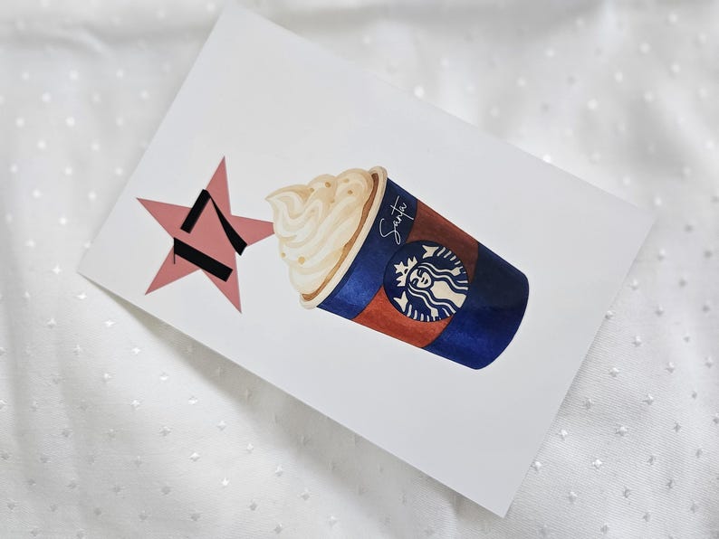 May include: Watercolor illustration of a Starbucks coffee cup with whipped cream, a pink star, and the word "Santa" written on the cup. The cup is brown and blue with the Starbucks logo.