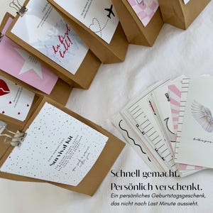 Op de afbeelding: Een verzameling bruine papieren zakken met witte kaarten bevestigd door zilveren metalen clips, met diverse grafische ontwerpen waaronder een gebroken hart, een ster en de tekst Survival Kit, naast een stapel witte wenskaarten met engelvleugels.