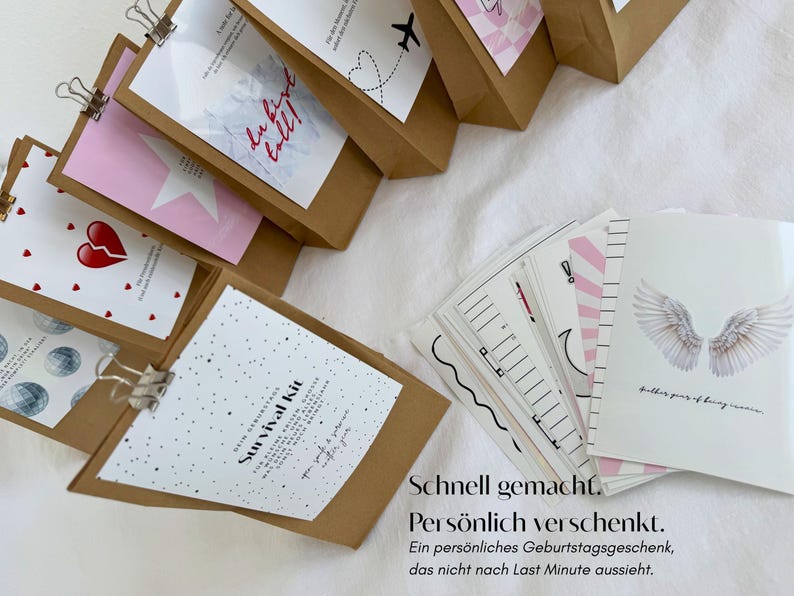 Op de afbeelding: Een verzameling bruine papieren zakken met witte kaarten bevestigd door zilveren metalen clips, met diverse grafische ontwerpen waaronder een gebroken hart, een ster en de tekst Survival Kit, naast een stapel witte wenskaarten met engelvleugels.