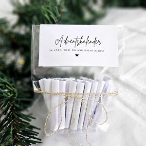May include: A clear cellophane bag tied with gold twine, containing 24 rolled white papers. A white card with black script reads "Adventskalender" and "24 Lose, weil du mir wichtig bist" with a heart symbol. Christmas tree branches are in the background.