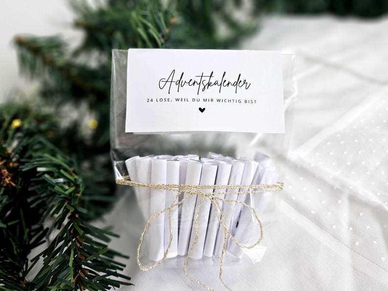 May include: A clear cellophane bag tied with gold twine, containing 24 rolled white papers. A white card with black script reads "Adventskalender" and "24 Lose, weil du mir wichtig bist" with a heart symbol. Christmas tree branches are in the background.