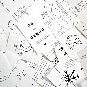 May include: A collection of white cards with black text and illustrations. The cards feature inspirational quotes, drawings of a crescent moon, stars, snowflakes, and angel wings. The cards are scattered across a surface.