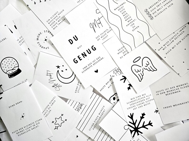 May include: A collection of white cards with black text and illustrations. The cards feature inspirational quotes, drawings of a crescent moon, stars, snowflakes, and angel wings. The cards are scattered across a surface.