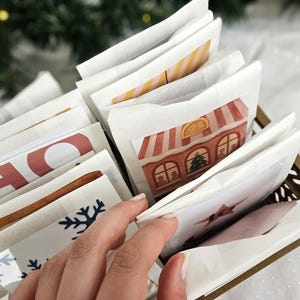 May include: A decorative advent calendar with individual paper packets, some with holiday-themed illustrations and text. The packets are arranged in a gold-colored basket. The image includes a hand reaching for one of the packets.