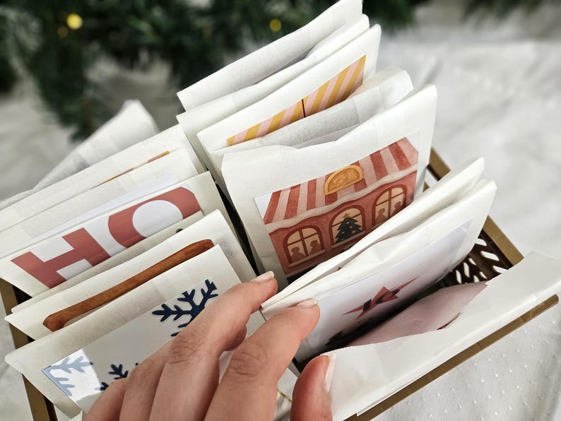 May include: A decorative advent calendar with individual paper packets, some with holiday-themed illustrations and text. The packets are arranged in a gold-colored basket. The image includes a hand reaching for one of the packets.