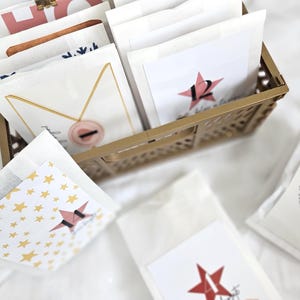 May include: A gold-colored metal basket filled with white paper bags. Some bags have a red star with a number on them. Other bags have gold star patterns or decorative designs. The theme is a holiday advent calendar.