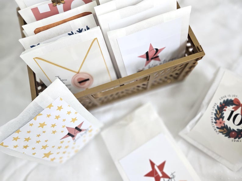 May include: A gold-colored metal basket filled with white paper bags. Some bags have a red star with a number on them. Other bags have gold star patterns or decorative designs. The theme is a holiday advent calendar.
