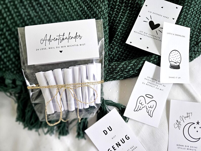 May include: A clear cellophane bag contains 24 rolled white papers tied with gold twine, labeled "Adventskalender." Several small white cards with black illustrations and text are scattered around, including a heart, snow globe, and angel wings.