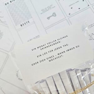 K&ouml;nnte beinhalten: Eine transparente Plastikt&uuml;te mit gerollten wei&szlig;en Papierstreifen, mit einem goldfarbenen Band am Boden. Eine wei&szlig;e Karte mit schwarzem Text befindet sich vor der T&uuml;te. Der Text lautet: "Ein Monat voller kleiner Erinnerungen: Ein Los f&uuml;r jeden Tag."