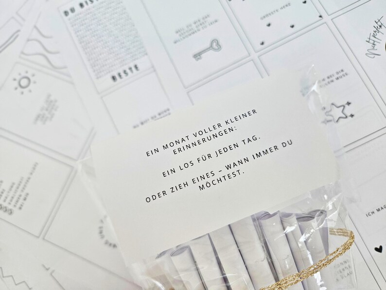 K&ouml;nnte beinhalten: Eine transparente Plastikt&uuml;te mit gerollten wei&szlig;en Papierstreifen, mit einem goldfarbenen Band am Boden. Eine wei&szlig;e Karte mit schwarzem Text befindet sich vor der T&uuml;te. Der Text lautet: "Ein Monat voller kleiner Erinnerungen: Ein Los f&uuml;r jeden Tag."