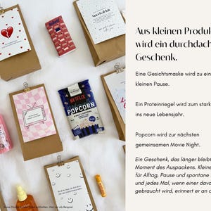 Op de afbeelding: Een verzameling bruine papieren cadeauzakjes met diverse artikelen, waaronder een zak Netflix-popcorn, een roze gezichtsmasker, een nude oogschaduwpalet, een eiwitreep en kaarten met tekst en illustraties, met Duitse tekst aan de rechterkant.