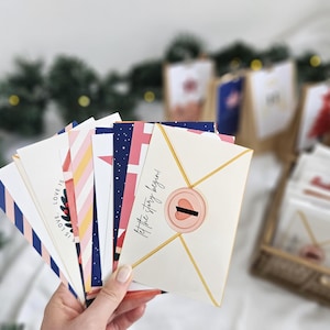 May include: A hand holds a collection of decorative envelopes with various patterns and text. The envelopes feature designs in navy, pink, white, and gold. One envelope reads "Let the story begin!" and has a pink seal.