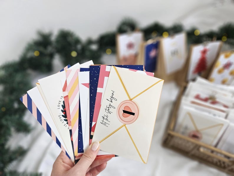 May include: A hand holds a collection of decorative envelopes with various patterns and text. The envelopes feature designs in navy, pink, white, and gold. One envelope reads "Let the story begin!" and has a pink seal.