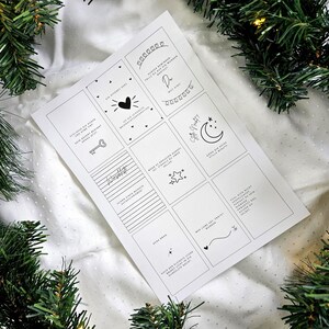 May include: A white paper sheet with multiple small, square designs, each containing a simple black illustration and text. The sheet is surrounded by green Christmas tree branches and lights.