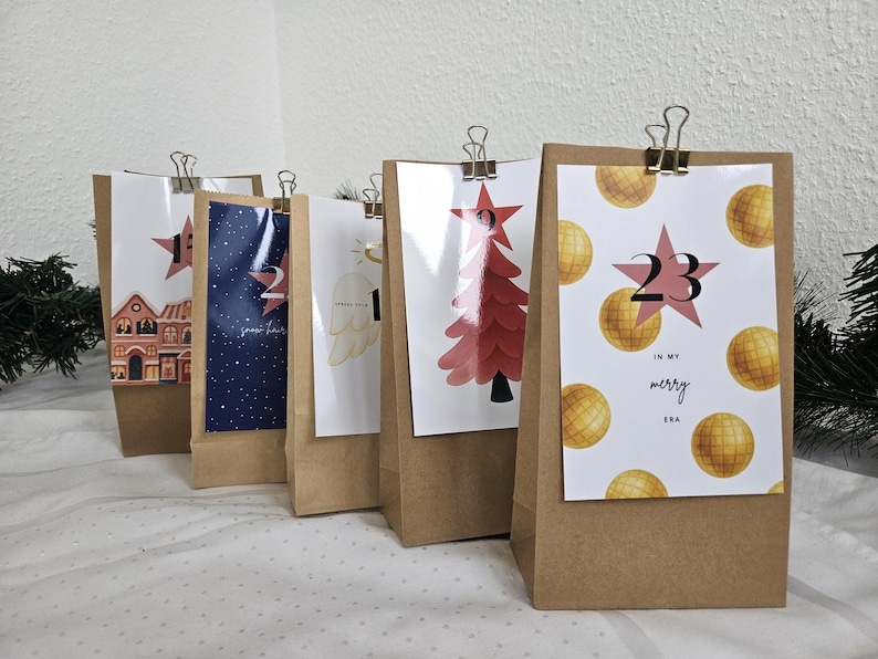 May include: Advent calendar with brown paper bags, each featuring a different festive design. The designs include a house, a starry night, an angel wing, a Christmas tree, and the number 23 with the words "in my merry era."
