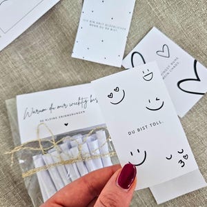 K&ouml;nnte beinhalten: Eine Sammlung wei&szlig;er Karten mit schwarzem Text und Illustrationen. Eine Karte lautet "DU BIST TOLL." mit Smileys. Eine andere Karte lautet "Warum du mir wichtig bis." Eine Hand h&auml;lt eine Karte mit rotem Nagellack.