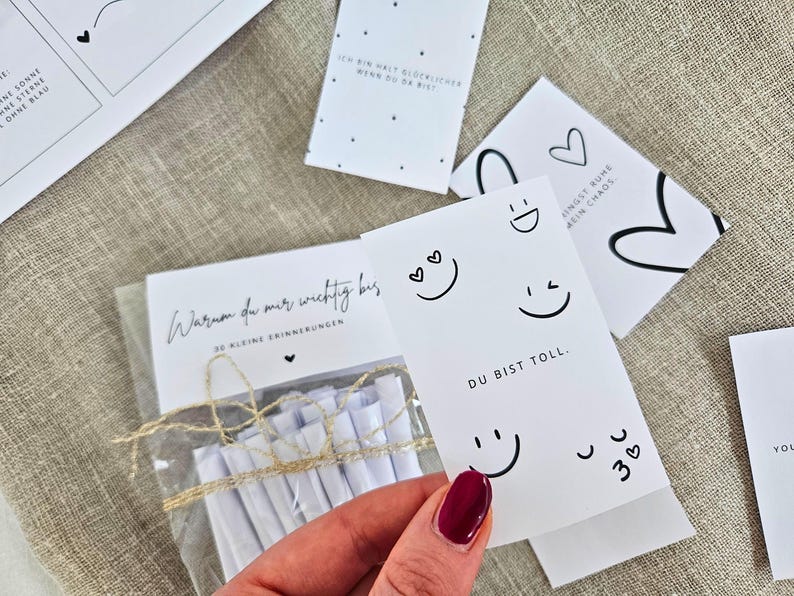 K&ouml;nnte beinhalten: Eine Sammlung wei&szlig;er Karten mit schwarzem Text und Illustrationen. Eine Karte lautet "DU BIST TOLL." mit Smileys. Eine andere Karte lautet "Warum du mir wichtig bis." Eine Hand h&auml;lt eine Karte mit rotem Nagellack.