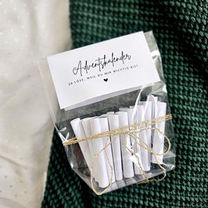 May include: A clear cellophane bag tied with gold twine, containing rolled white paper scrolls. A white card with black script reads "Adventskalender" and "24 Lose. Weil du mir wichtig bist" with a heart symbol.