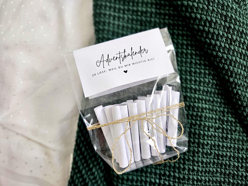 May include: A clear cellophane bag tied with gold twine, containing rolled white paper scrolls. A white card with black script reads "Adventskalender" and "24 Lose. Weil du mir wichtig bist" with a heart symbol.