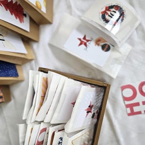 May include: A collection of small paper bags and packets, likely for a holiday advent calendar. The bags are brown and white, with various illustrations and numbers. Some packets have star and wreath designs. A festive "HO HO HO" sign is visible.