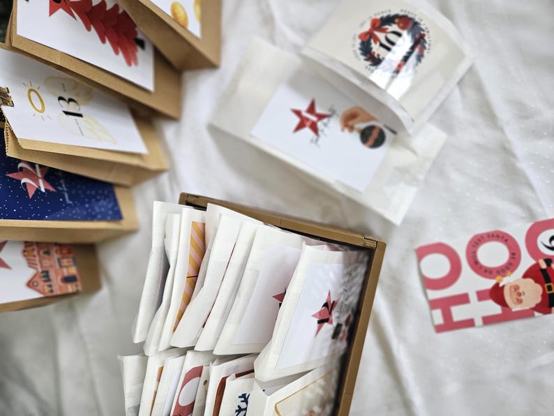 May include: A collection of small paper bags and packets, likely for a holiday advent calendar. The bags are brown and white, with various illustrations and numbers. Some packets have star and wreath designs. A festive "HO HO HO" sign is visible.