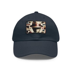 Puede incluir: Gorra de béisbol azul marino con un parche rectangular. El parche muestra una ilustración vintage de un sombrero fedora sobre una cabeza de maniquí, rodeada de varios diseños de sombreros. La gorra tiene una visera curva.