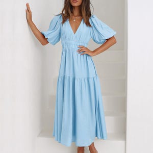 K&ouml;nnte beinhalten: Ein hellblaues Midikleid mit V-Ausschnitt und kurzen Puff&auml;rmeln. Das Kleid hat eine taillierte Taille und einen Stufenrock. Das Model tr&auml;gt nudefarbene Sandalen. Das Kleid ist aus einem weichen, flie&szlig;enden Stoff gefertigt.