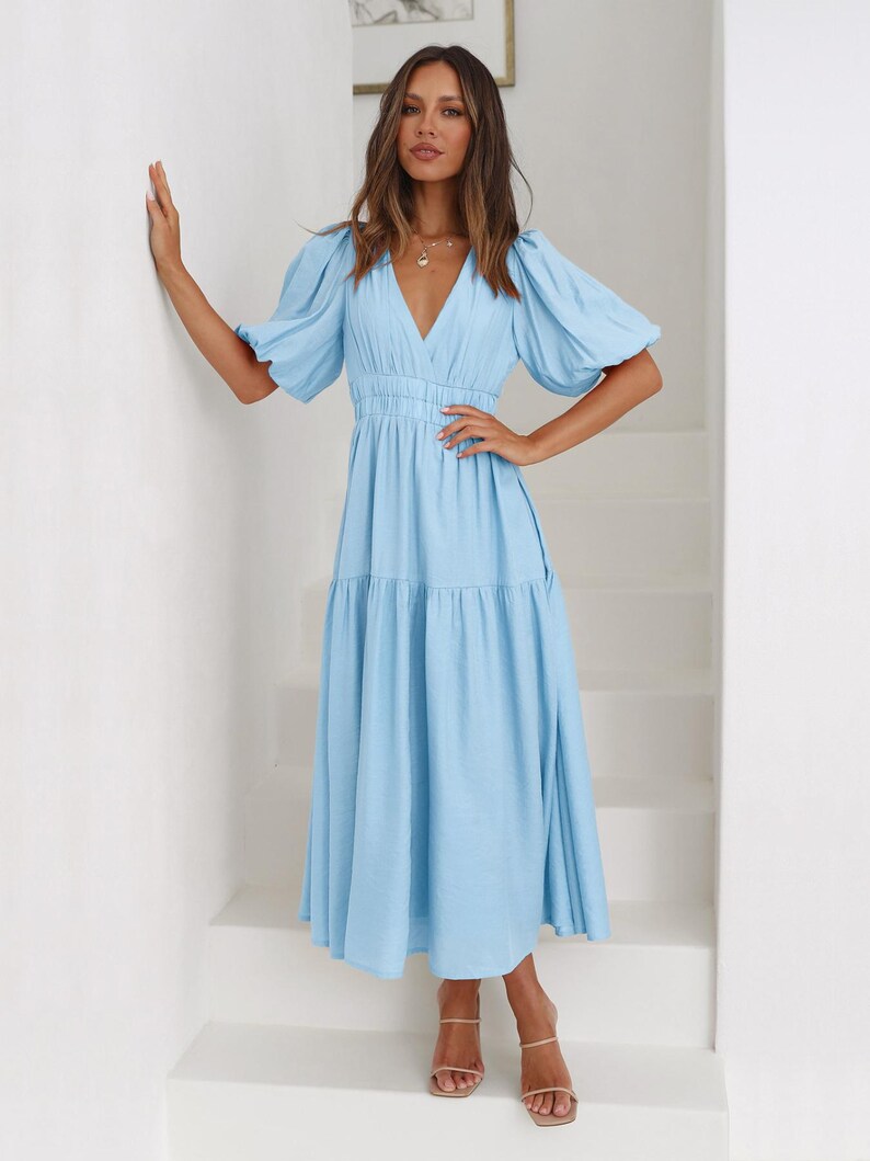 K&ouml;nnte beinhalten: Ein hellblaues Midikleid mit V-Ausschnitt und kurzen Puff&auml;rmeln. Das Kleid hat eine taillierte Taille und einen Stufenrock. Das Model tr&auml;gt nudefarbene Sandalen. Das Kleid ist aus einem weichen, flie&szlig;enden Stoff gefertigt.