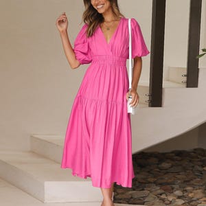 K&ouml;nnte beinhalten: Ein leuchtend pinkfarbenes Midikleid mit kurzen, gepufften &Auml;rmeln und einem V-Ausschnitt. Das Kleid hat eine taillierte Taille und einen Stufenrock. Die Person tr&auml;gt wei&szlig;e Sandalen und eine wei&szlig;e Umh&auml;ngetasche. Der Hintergrund zeigt eine wei&szlig;e Treppe.