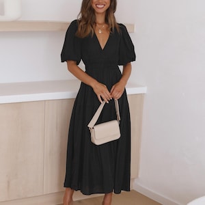 K&ouml;nnte beinhalten: Ein schwarzes Midikleid mit V-Ausschnitt und kurzen Puff&auml;rmeln. Das Kleid hat eine taillierte Taille und einen flie&szlig;enden Rock. Eine kleine, cremefarbene Handtasche mit Schulterriemen wird gehalten. Beigefarbene Abs&auml;tze vervollst&auml;ndigen den Look.